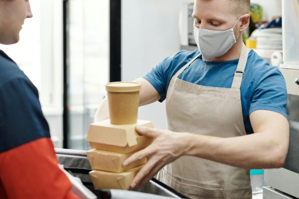 Masked worker packs takeaway food for delivery in restaurant kitchen.